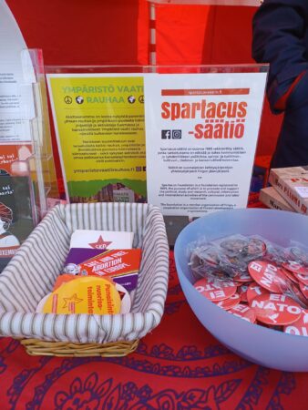 Teltan pöydällä astioissa rintamerkkejä ja tarroja sekä Ympäristö vaatii rauhaa -hankkeen ja Spartacus-säätiön esittelytaulut.