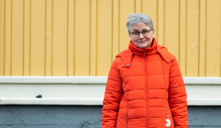 Mervi Grönfors seisoo keltaisen puutalon seinustalla.