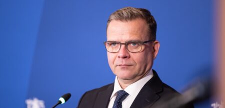 Petteri Orpo katsoo kameraan sinisen taustan edessä.