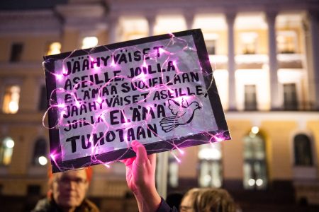 Kädessä pidetty kyltti mielenosoituksessa iltahämärässä. Kyltissä lukee ”Jäähyväiset aseille, joilla elämää suojellaan. Jäähyväiset aseille, joilla elämä tuhotaan” ja siinä on rauhankyyhkyn kuva. Kyltin ympärille on kiedottu vaaleanpunaiset valonauhat. Taustalla näkyy valaistu rakennus ja mielenosoittajia.