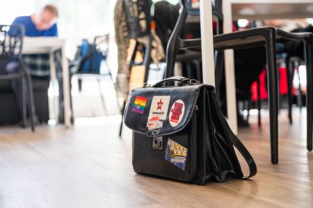 Musta nahkainen salkku lattialla pöytien ja tuolien keskellä sisätilassa. Salkussa on poliittisia tarroja, muun muassa sateenkaarilippu, kommunistien tähti ja Euroopan vasemmiston tarra ”We want direct democracy now”.