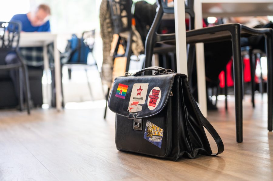 Musta nahkainen salkku lattialla pöytien ja tuolien keskellä sisätilassa. Salkussa on poliittisia tarroja, muun muassa sateenkaarilippu, kommunistien tähti ja Euroopan vasemmiston tarra ”We want direct democracy now”.