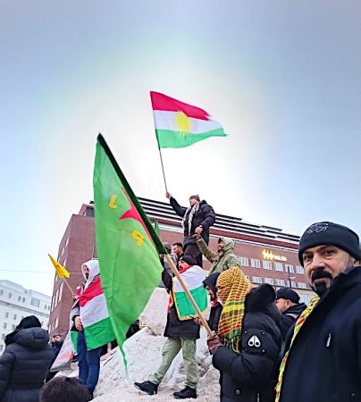 Ulkona järjestetty mielenosoitus Helsingin keskustassa Kampissa. Ryhmä ihmisiä seisoo lumisella korokkeella ja pitelee kurdilippuja. Yksi henkilö nostaa lipun korkealle ilmaan. Osallistujilla on talvivaatteet ja huiveja, ja taustalla näkyy suuria rakennuksia.