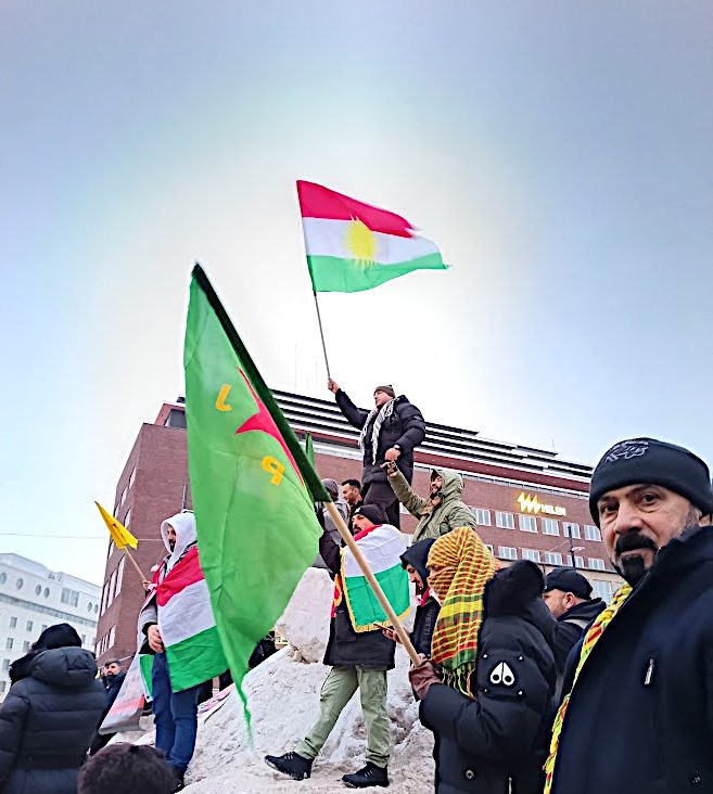 Ulkona järjestetty mielenosoitus Helsingin keskustassa Kampissa. Ryhmä ihmisiä seisoo lumisella korokkeella ja pitelee kurdilippuja. Yksi henkilö nostaa lipun korkealle ilmaan. Osallistujilla on talvivaatteet ja huiveja, ja taustalla näkyy suuria rakennuksia.